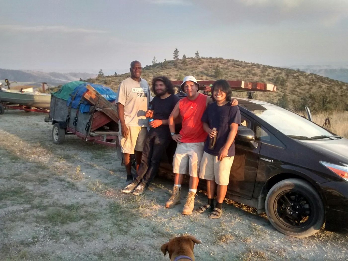 A hatchback towing a trailer loaded with salvaged lumber on a dirt track in the hills, with four people standing beside it.
